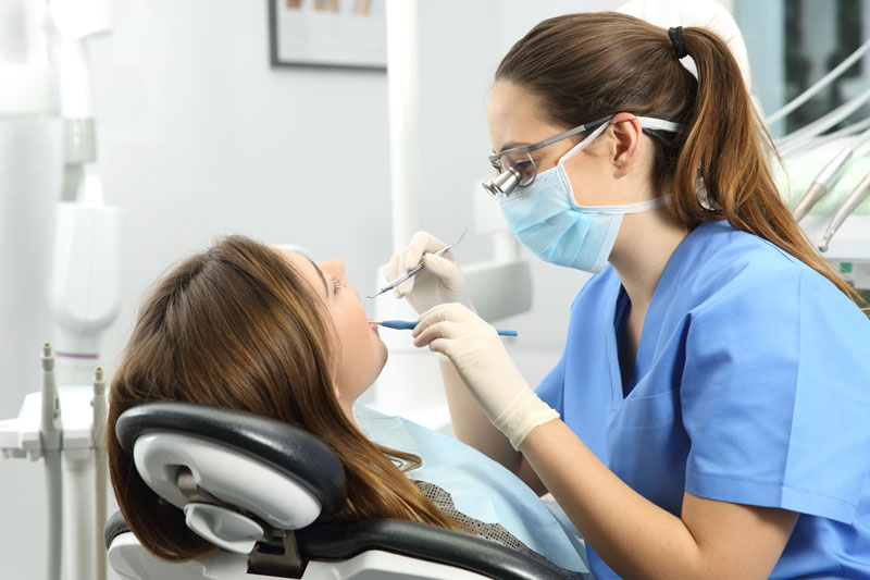 A dentist, wearing a blue surgical mask and gloves, is examining a patient's teeth with dental instruments. The patient is reclined in a dental chair, and the dental office is equipped with modern tools and equipment.